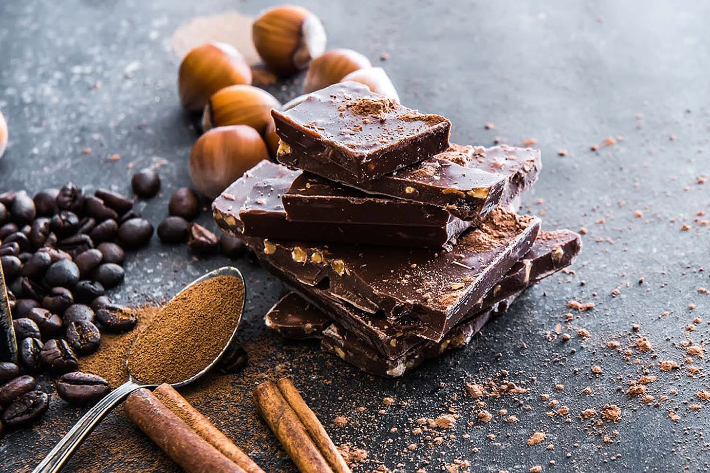 Broken up bars of chocolate rest on slate with a spoonful of cocoa powder and coffee beans and hazelnuts around.